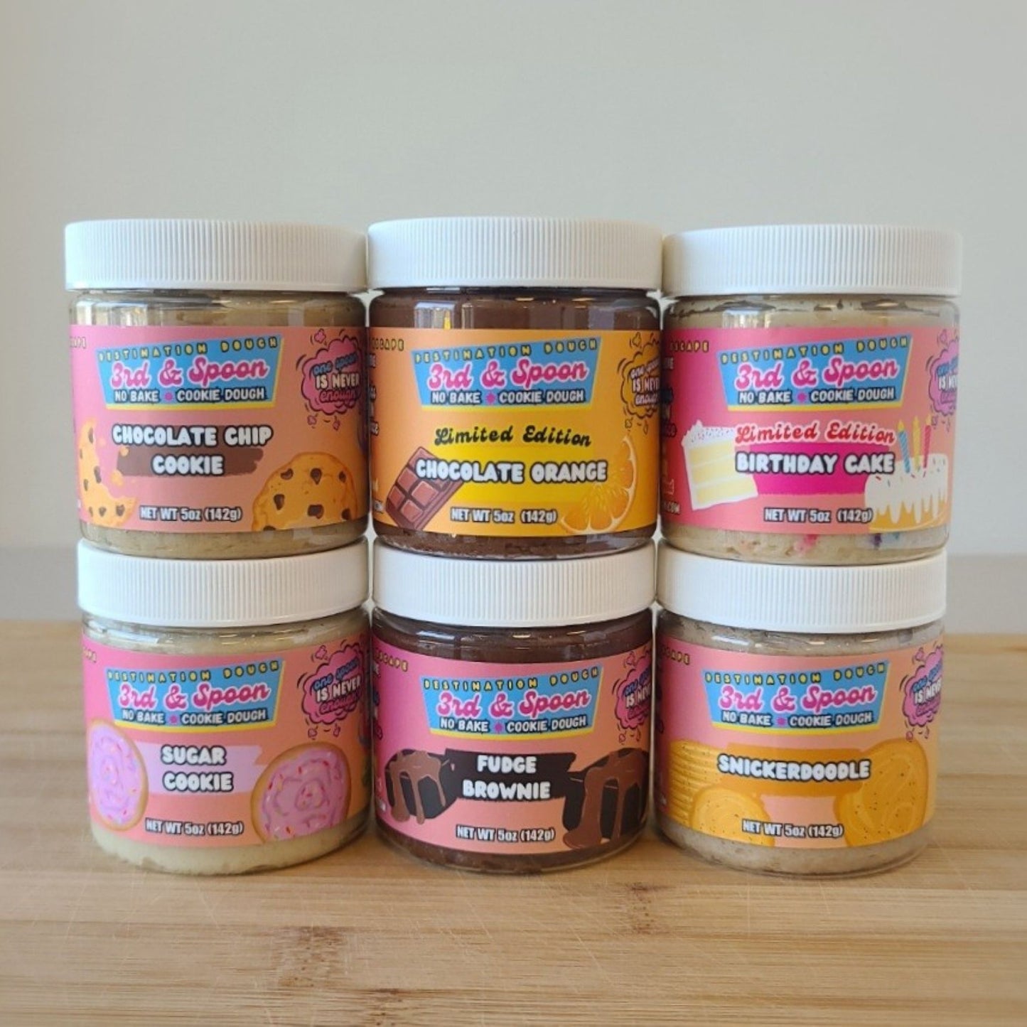 Six 5oz jars of 3rd & Spoon cookie dough on a wooden surface with colorful labels.